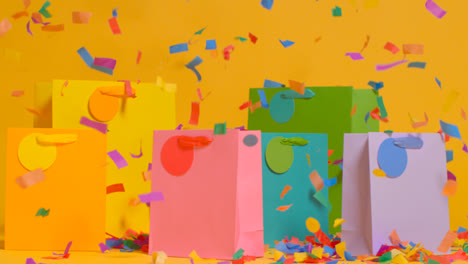 Studio-Shot-Of-Colourful-Birthday-Party-Gift-Bags-Against-Yellow-Background-With-Falling-Paper-Confetti-2